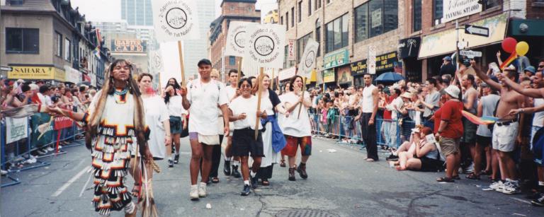 Un groupe de personnes portant des pancartes sur lesquelles on peut lire « 2-Spirited People of the 1st Nations » marchent au centre d’une rue de la ville, tandis que des gens regardent derrière des barrières de part et d’autre de la rue. Visibilité masquée.