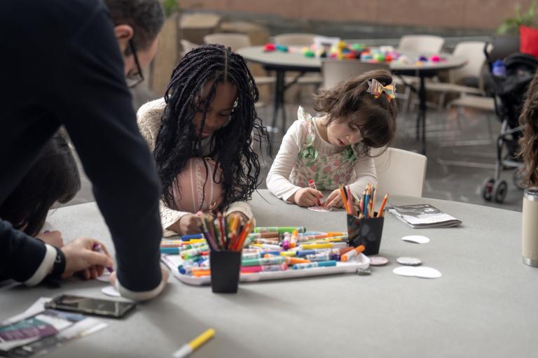 Deux enfants sont assis à une table et font de l’art tandis qu'un adulte s'appuie sur la table pour les observer. Visibilité masquée.