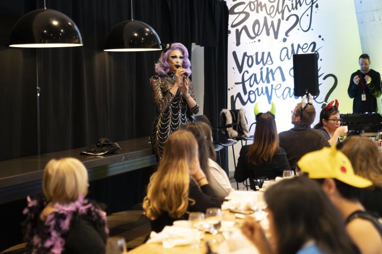 Une artiste de drag aux cheveux violets et à la robe argentée et noire s'exprime devant un groupe qui brunch. Visibilité masquée.