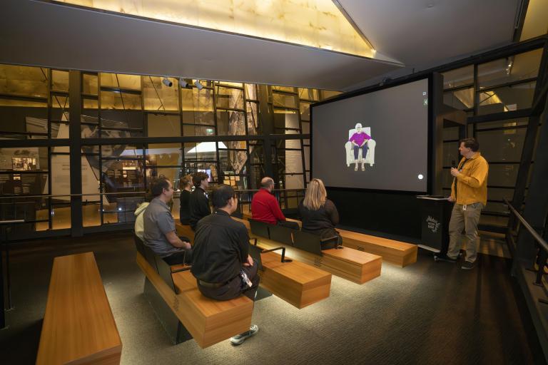 Un petit groupe de personnes assises sur des bancs en bois dans un théâtre fait face à un écran montrant une femme âgée assise dans un fauteuil. Un homme vêtu d’une chemise jaune se tient à droite de l’écran et tient un microphone. Visibilité masquée.