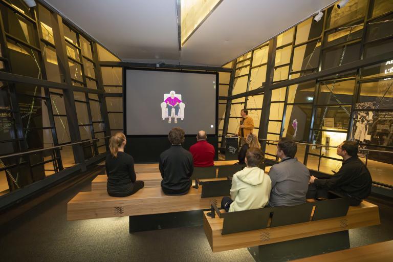 Un petit groupe de personnes assises sur des bancs en bois dans un théâtre fait face à un écran montrant une femme âgée assise dans un fauteuil. Un homme vêtu d’une chemise jaune se tient à droite de l’écran et tient un microphone. Visibilité masquée.