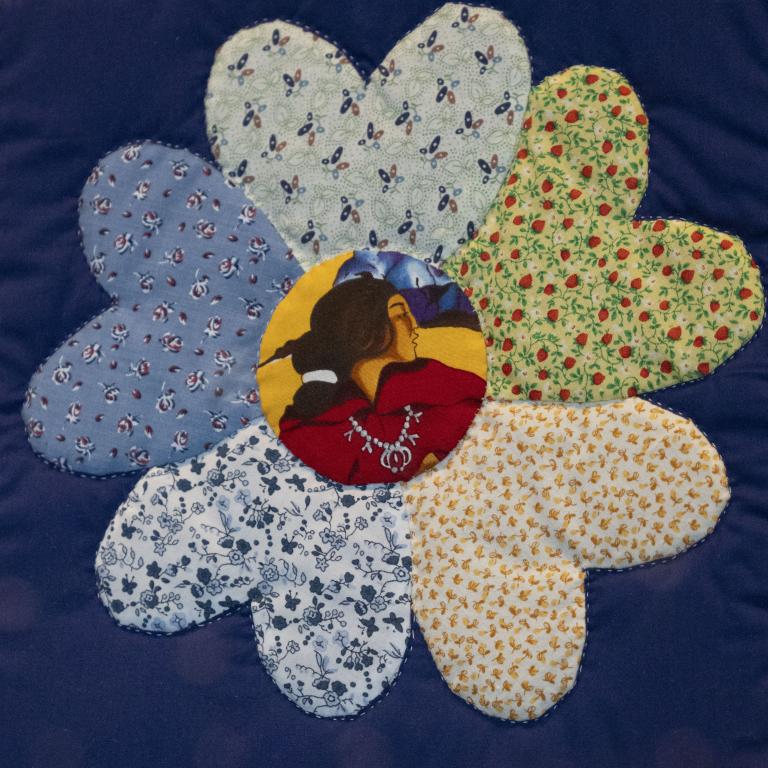 Gros plan sur une fleur sur une courtepointe. Chacun des pétales est un tissu fleuri différent, tandis qu’au centre de la fleur se trouve un tissu sur lequel est imprimé le profil d’une femme autochtone. Visibilité masquée.