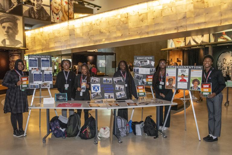 Dans une salle de musée, six étudiants noirs présentent fièrement leur travail sur des affiches et sur une table. Visibilité masquée.