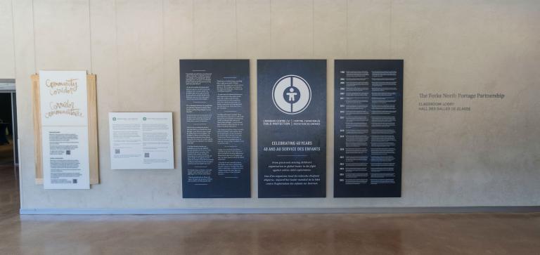 Grands panneaux bleu foncé avec texte et graphiques blancs exposés dans le Corridor communautaire du Musée canadien pour les droits de la personne. Le panneau central comporte un logo circulaire et commémore le 40e anniversaire du Centre canadien de protection de l'enfance. Les deux panneaux de chaque côté présentent du texte bilingue en anglais et en français. Visibilité masquée.