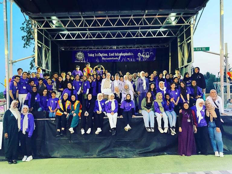 Des dizaines de personnes sont rassemblées sur une scène, la plupart souriantes et vêtues de violet. Beaucoup portent le hidjab. Une banderole est suspendue à l’arrière de la scène sur laquelle on peut lire, en anglais « Prenons l’initiative. Mettons fin à l’islamophobie. #OurLondonFamily ». Visibilité masquée.