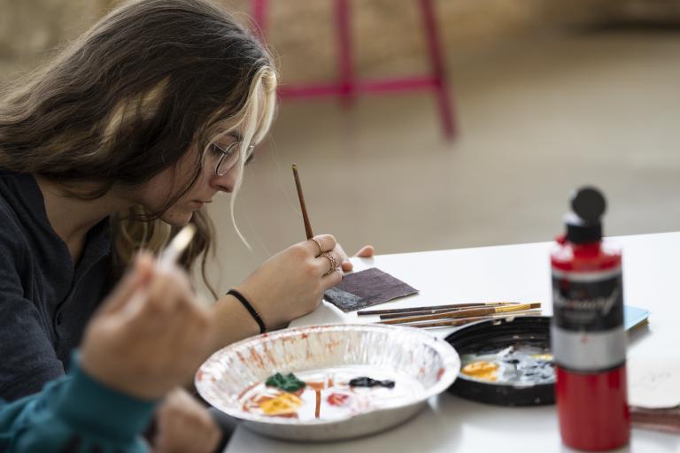 A young woman sitting at a table paints a design on a tile while a tin tray with various paints sits in front of her. An open bottle of red paint sits on the table. Visibilité masquée.