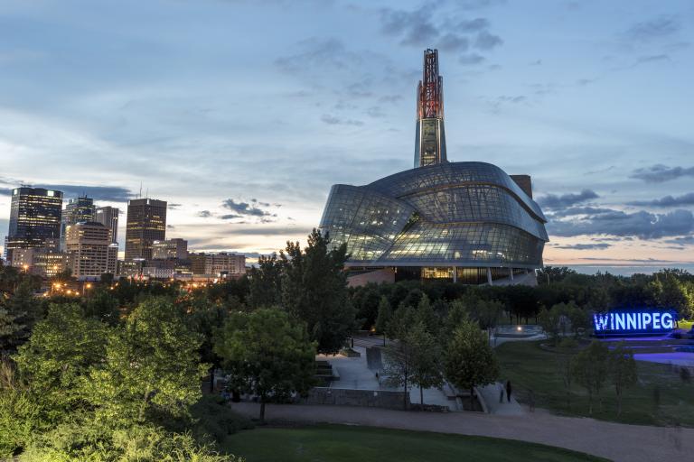 Photo extérieure des fenêtres en verre et de la tour du Musée canadien pour les droits de la personne, prise au crépuscule avec le ciel des prairies en arrière-plan. Le panneau Winnipeg à Forks est situé à droite de l'image. Plusieurs tours de bureaux et les lumières du centre-ville sont visibles à gauche de l'image. Visibilité masquée.