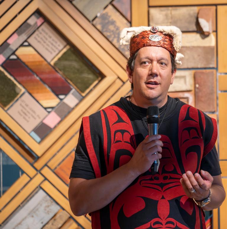 Un homme debout, micro à la main, s’adresse à une foule vêtue de costumes traditionnels autochtones devant une installation artistique en panneaux de bois. Visibilité masquée.
