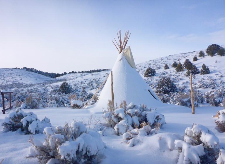 Un tipi recouvert de neige se dresse dans un paysage vallonné parsemé d’arbres et de buissons, sous un ciel bleu. Visibilité masquée.