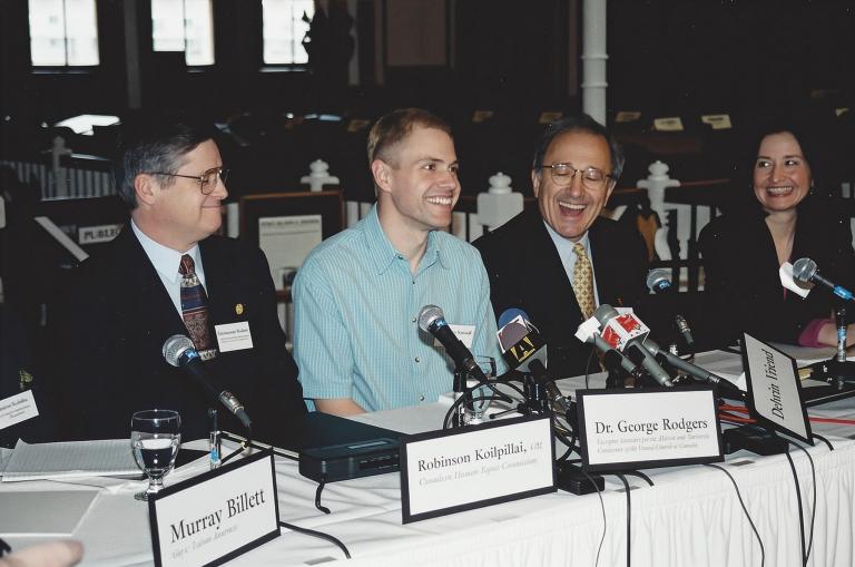 Un homme souriant vêtu d’une chemise bleu clair est assis à une longue table devant un groupe de microphones. Assis de part et d’autre de lui, d’autres personnes en complet sont également tournées vers les microphones et sourient. Les plaquettes nominatives le long de la table indiquent, de gauche à droite, Murray Billet, Robinson Koilpillai et Dr George Rodgers, avec des affiliations illisibles sous les noms. Une autre plaquette, renversée sur le côté, indique Delwin Vriend sans affiliation. Visibilité masquée.