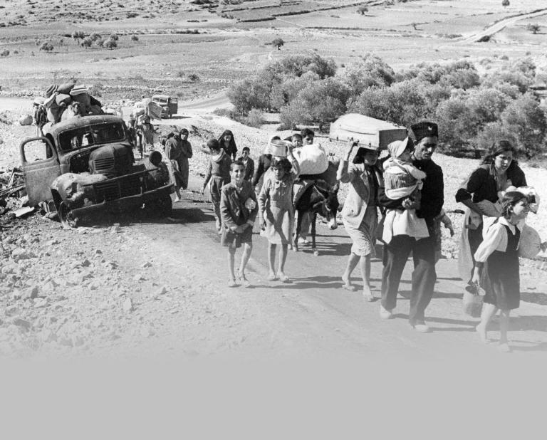 Une photo en noir et blanc d’un groupe d’adultes et d’enfants transportant leurs affaires, marchant sur une route désertique non pavée près d’un camion en ruine. Visibilité masquée.