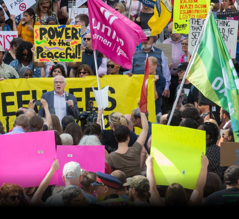 Un homme blanc aux cheveux blancs, portant des lunettes et un blazer, parle au micro. Il est entouré d'une foule de personnes brandissant des appareils photo, des pancartes de protestation et une banderole. Visibilité masquée.