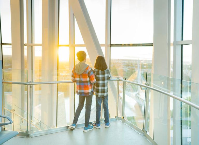 Deux enfants se tiennent dans une tour, dos à la caméra, et regardent vers l’horizon qui surplombe un paysage urbain. Visibilité masquée.