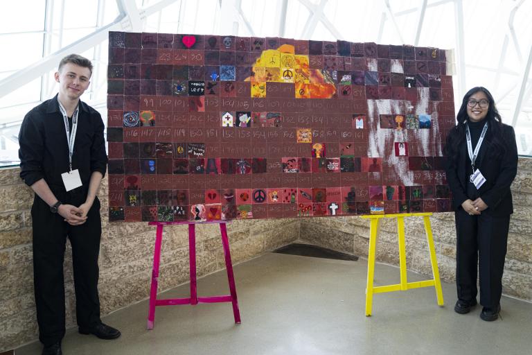 Deux élèves debout de chaque côté d’une grande œuvre d’art exposée sur deux chevalets. Visibilité masquée.