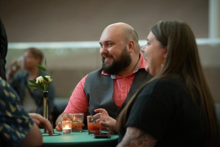 Un homme et une femme sont assis ensemble à une table, souriant chaleureusement tout en écoutant d’autres convives. L’homme porte une chemise saumon et un gilet gris, et de petites bougies et des boissons sont disposées sur la table, créant une atmosphère intime et chaleureuse. Visibilité masquée.