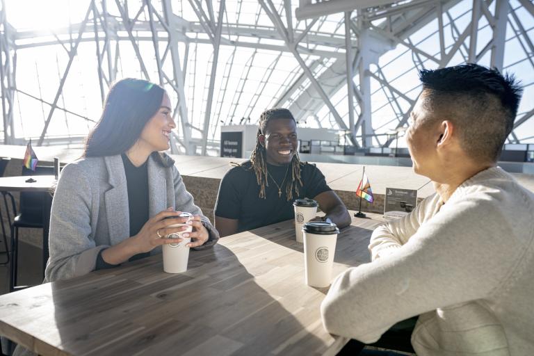 Trois personnes sont assises à une table. Elles sourient toutes, un café devant elles. Un petit drapeau de la Fierté progressif est posé sur leur table et sur la table vide derrière elles. Le soleil brille à travers les fenêtres du Musée. Visibilité masquée.