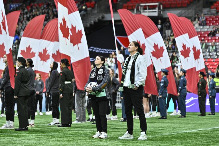 Des personnes se tiennent debout sur le terrain d’un stade pendant une cérémonie d’avant-match, tandis que de grands drapeaux canadiens sont brandis derrière elles, remplissant l’arrière-plan de rouge et de blanc. Visibilité masquée.