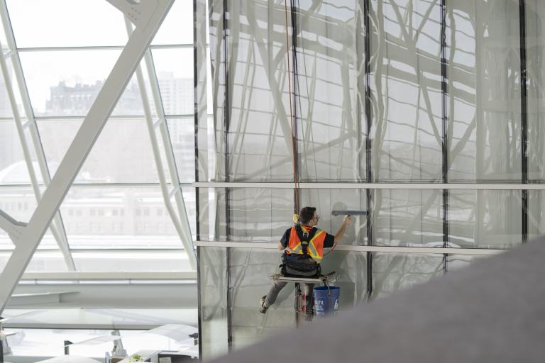 Un travailleur portant un gilet de sécurité et suspendu à des cordes nettoie des fenêtres en hauteur. Visibilité masquée.