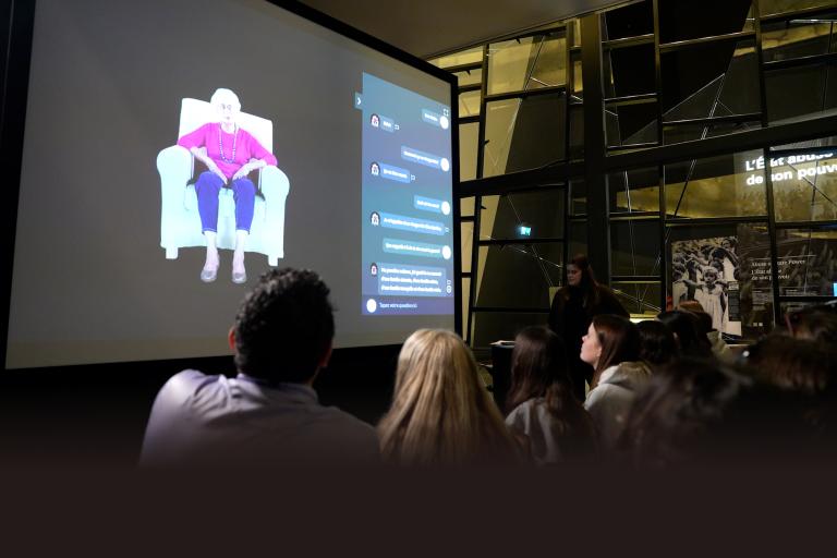Un auditoire regarde un grand écran de projection sur lequel apparaît une femme âgée vêtue d’un haut violet et d’un pantalon bleu, assise dans un fauteuil blanc, à côté d’une interface interactive. La présentation se déroule dans un espace moderne à l’architecture géométrique en verre, et une personne se tient à droite de l’écran. Visibilité masquée.