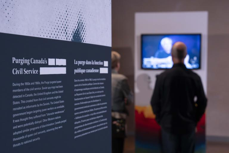 Un panneau d’exposition portant le texte « La purge dans la fonction publique canadienne » se trouve à gauche. Deux personnes à l’arrière-plan, à droite, regardent une vidéo.