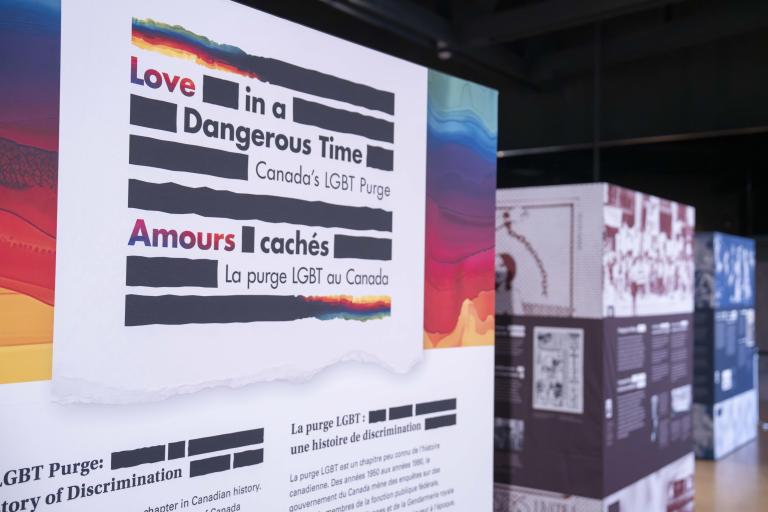 Une grande affiche est composée de lignes horizontales noires épaisses et les mots « Love in a Dangerous Time: Canada’s LGBT Purge » en anglais et « Amours cachés : La purge LGBT au Canada » en français sont répartis à l’intérieur des lignes. Les lignes du haut et du bas sont un mélange de noir et de couleurs arc-en-ciel.