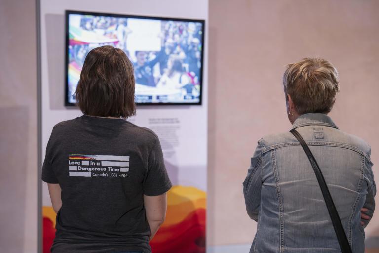 Deux personnes nous tournent le dos et regardent une vidéo. Le mur sur lequel est accroché le moniteur vidéo est décoré de couleurs arc-en-ciel.