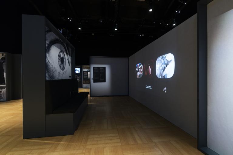Un panneau d’exposition à gauche présente l’image d’un grand œil. À droite, trois petites vidéos sont projetées sur le mur. L’espace est sombre.