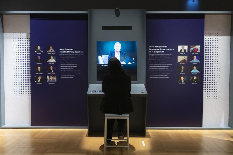 Une personne nous tournant le dos regarde une vidéo. De petits portraits de personnes encadrent le moniteur vidéo à gauche et à droite.