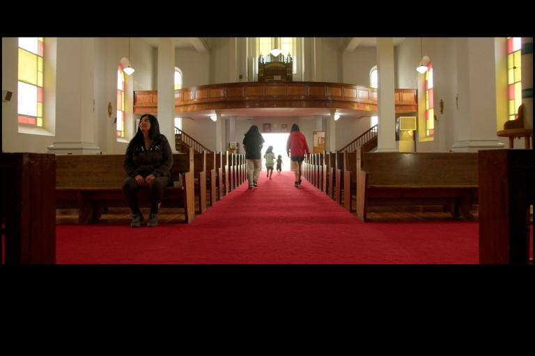 Intérieur d’une église avec une longue allée recouverte d’un tapis rouge entre des bancs en bois. La lumière du soleil filtre à travers les vitraux tandis que plusieurs enfants courent vers l’entrée lumineuse au fond de l’église et qu’une femme autochtone est assise seule sur un banc à gauche. Visibilité masquée.