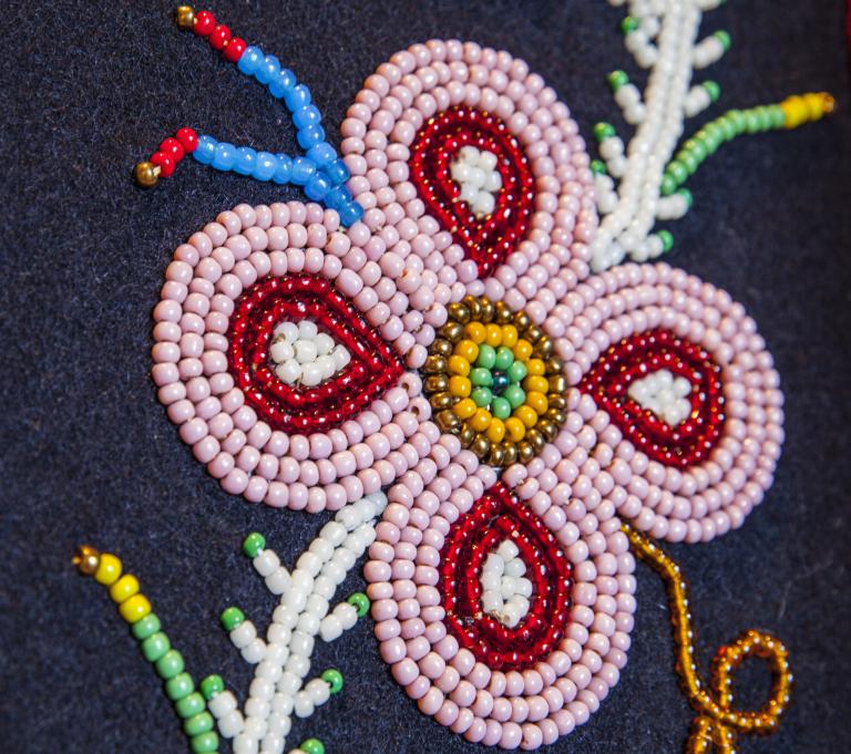 Gros plan sur un motif floral perlé sur un tissu foncé, représentant une fleur rose à quatre pétales avec des détails rouges et blancs, un centre perlé jaune et vert, et des perles tout autour ressemblant à des feuilles et des tiges. Visibilité masquée.