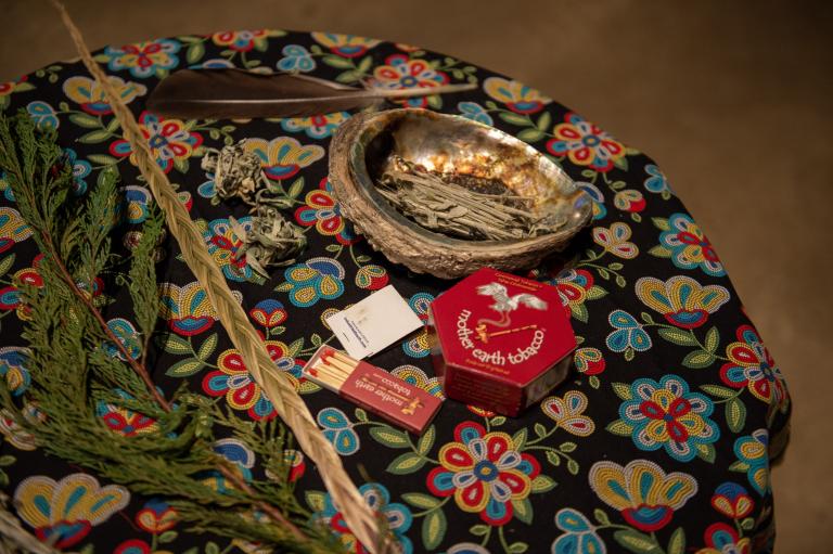 Des objets pour la purification disposés sur une table ronde recouverte d’une nappe à fleurs, notamment un coquillage abalone contenant de la sauge séchée, une plume, du foin d’odeur tressé, des branches de cèdre et une petite boîte d’allumettes. Visibilité masquée.