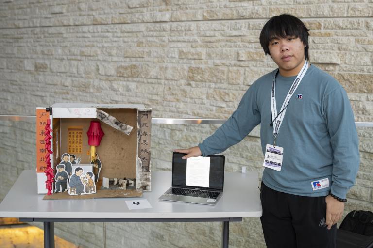 Un jeune homme se tient à côté d’une maquette ressemblant à un restaurant chinois. Un côté est bien rangé et comporte des illustrations de personnes en train de manger. L’autre côté est vide et endommagé.