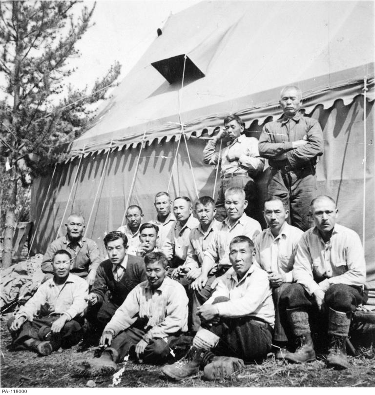 Un groupe d’hommes pose pour une photo avec une grande tente et un arbre en arrière-plan. Visibilité masquée.
