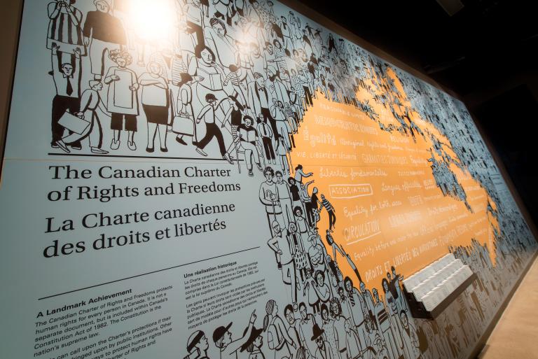 Sur le mur d’une exposition muséale figure une fresque illustrée représentant divers personnages et le titre « The Canadian Charter of Rights and Freedoms / La Charte canadienne des droits et libertés ». La fresque passe d’illustrations noires représentant des personnes à une partie orange où sont énoncés, en anglais et en français, les droits et libertés fondamentaux, qui prennent la forme du Canada. Visibilité masquée.