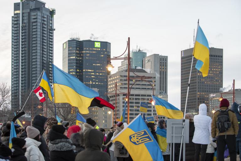 Une foule de personnes emmitouflées se tient debout dans une rue de ville pendant l’hiver, tenant de multiples drapeaux ukrainiens bleus et jaunes ainsi qu’un drapeau canadien. Le rassemblement semble être une manifestation en appui à l’Ukraine. De grands édifices à bureaux et des feux de circulation sont à l’arrière-plan sous un ciel couvert. Visibilité masquée.