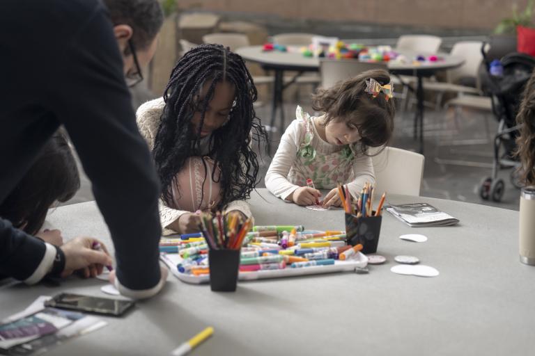 Deux jeunes enfants sont assis côte à côte à une table ronde, occupés à une activité artistique avec des marqueurs et des crayons de couleur. Visibilité masquée.