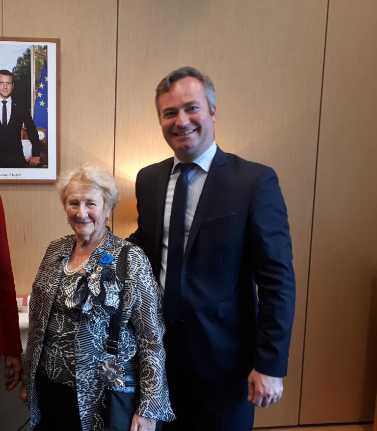 Une femme âgée aux cheveux courts et gris sourit doucement à la caméra. Elle porte des boucles d’oreilles en perles, un collier de perles et un chemisier à motifs noir et blanc. Elle se tient debout, baignée d’une lumière chaleureuse, devant un mur lambrissé. Visibilité masquée.