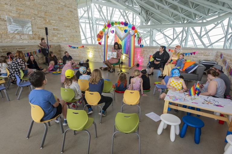 Une artiste drag fait la lecture à un groupe d'enfants et d'adultes assis sur des chaises colorées, dans un lieu lumineux de style industriel décoré de banderoles arc-en-ciel et de guirlandes, à l'occasion d'une heure du conte sur le thème de la Fierté. Visibilité masquée.