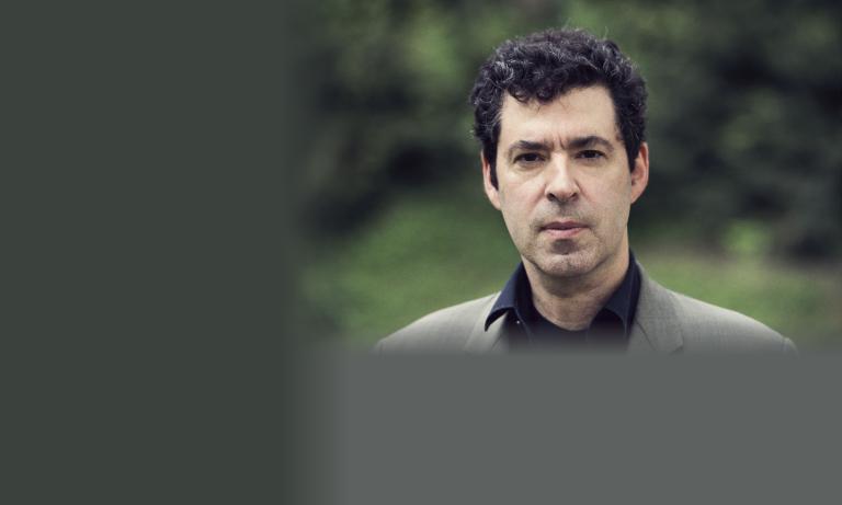 Portrait d’un homme d’âge mûr aux cheveux bruns bouclés, vêtu d’un blazer gris et d’une chemise noire, photographié en extérieur sur fond de feuillage vert flou. Son regard est sérieux et franc. Visibilité masquée.