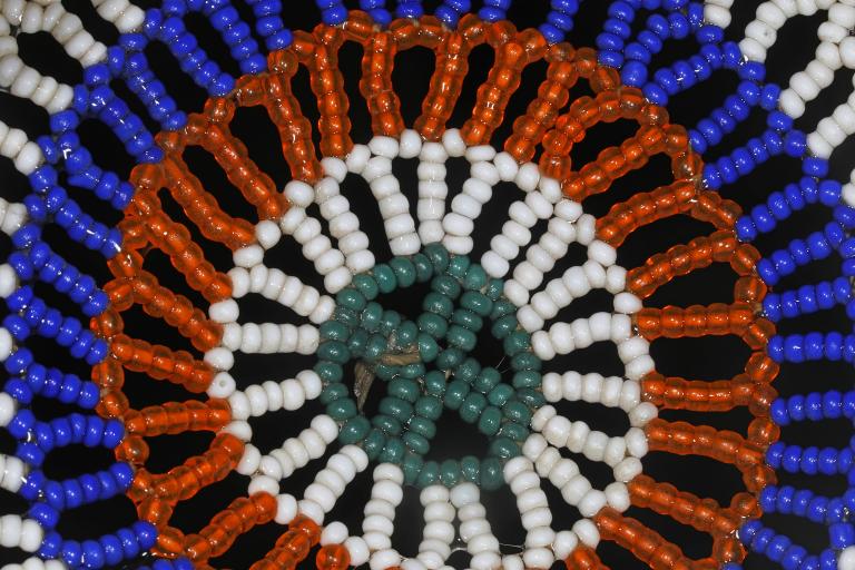 Une photo en gros plan d’un motif circulaire en perles sur fond noir. Le motif se compose d’anneaux concentriques de perles turquoise, blanches, orange et bleu vif, disposées en forme de fleur ou de soleil. Visibilité masquée.