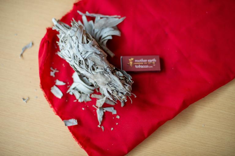 Un petit paquet de sauge séchée repose sur un tissu rouge vif, entouré de fragments de feuilles éparpillés. À côté se trouve une petite boîte d’allumettes rectangulaire portant l’inscription « Mother Earth Tobacco ». Ces objets sont posés sur une surface en bois clair. Visibilité masquée.
