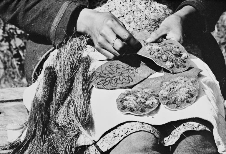 Une photo en noir et blanc montrant des mains tenant et cousant des morceaux de tissu décorés de broderies florales et de fibres texturées, étalés sur les genoux d’une personne. Visibilité masquée.