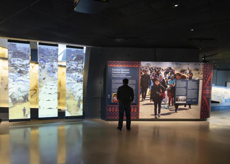Plan d’ensemble d'une galerie du Musée. À gauche, trois panneaux verticaux présentent une photo de tentes blanches et de personnes déplacées. À droite de l’image, une personne se tient debout devant un mur d’exposition et lit le texte. Visibilité masquée.