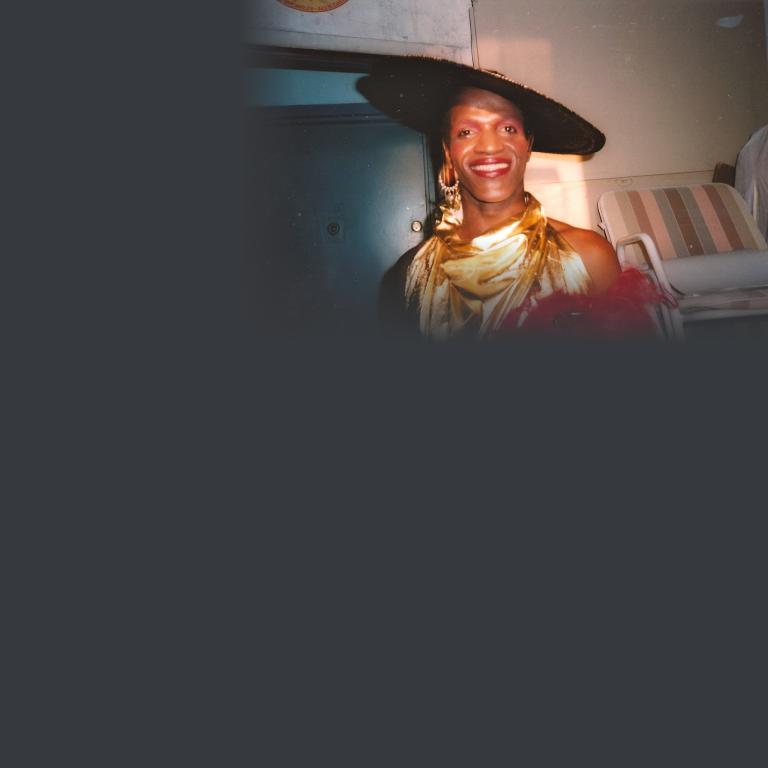 Une personne souriante vêtue d’une tenue de scène glamour, composée d’un chapeau noir à large bord orné de dentelle, d’un haut dos nu drapé en satin doré, d’accessoires à plumes rouges et de boucles d’oreilles en forme de lustres. La photo semble être un cliché vintage pris dans une loge en coulisses. Visibilité masquée.