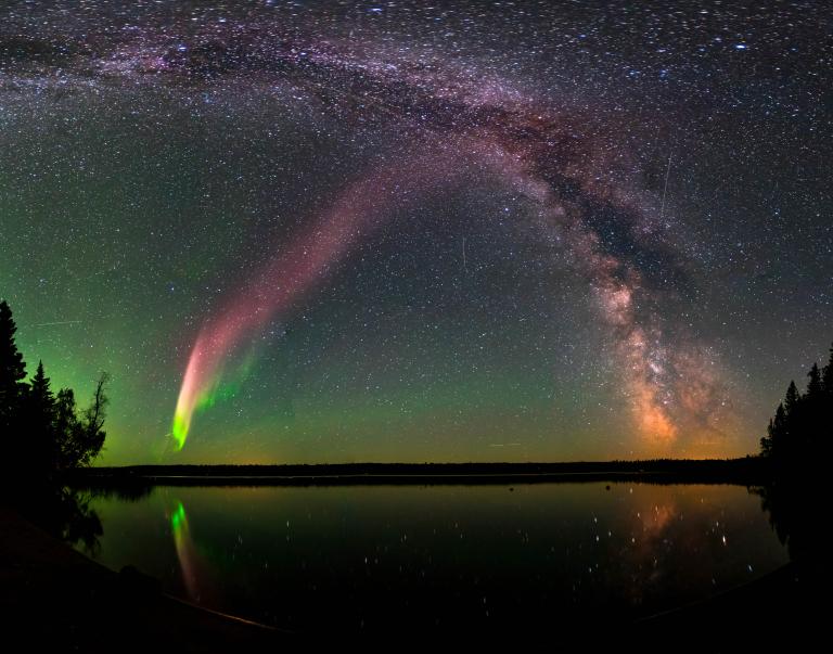 Un arc éclatant d’aurores rose-rougeâtre et vertes se dessine dans le ciel nocturne étoilé, le long du cœur de la Voie lactée, au-dessus d’un lac sombre bordé d’arbres dont la surface calme reflète ces lumières. Visibilité masquée.