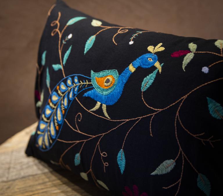 Photo en gros plan d’un coussin décoratif rectangulaire noir orné d’une broderie détaillée représentant un paon stylisé au cou bleu incurvé, à la crête bleu sarcelle et à la plume aux motifs oculaires orange et bleu sarcelle, posé sur un banc en bois. Le motif comprend également des surpiqûres marron en forme de vigne, des feuilles bleu sarcelle et sauge, ainsi que de petits motifs floraux bordeaux et blancs disséminés sur le tissu noir. Visibilité masquée.