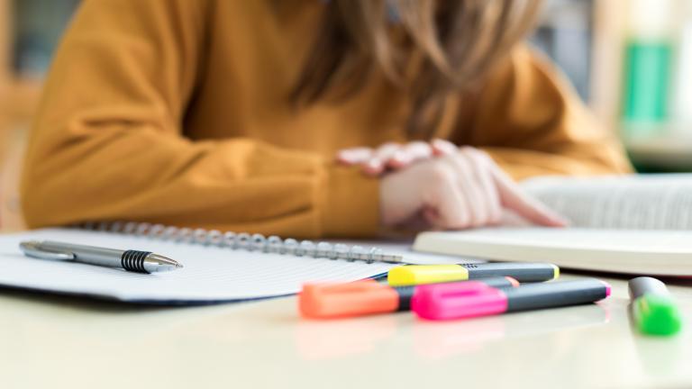 Gros plan sur un cahier à spirales, un stylo et des surligneurs colorés posés sur un bureau, avec, à l’arrière-plan, une personne vêtue d’un chandail couleur moutarde, dont la silhouette est floue. Visibilité masquée.