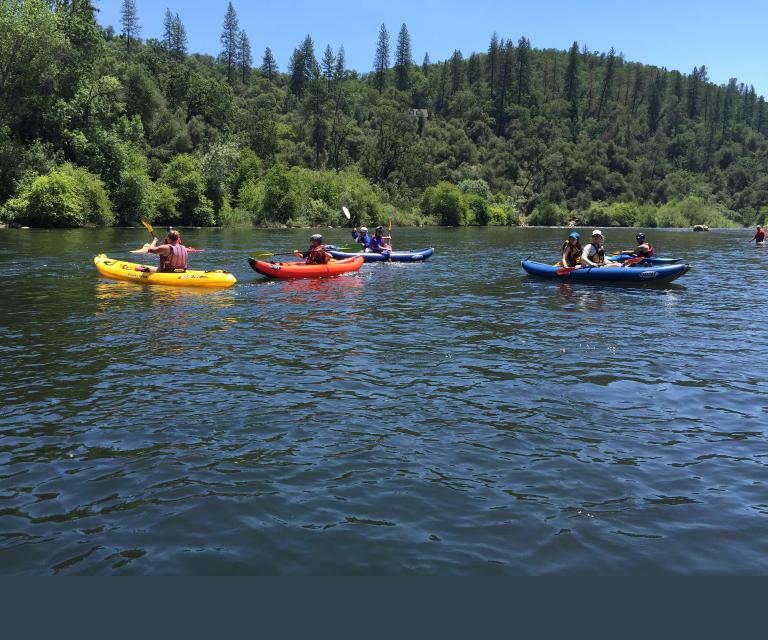 Des kayakistes pagayant dans des kayaks de couleurs jaune, orange et bleu sur une rivière calme, avec en arrière-plan une colline boisée de pins et de chênes sous un ciel bleu limpide. Visibilité masquée.