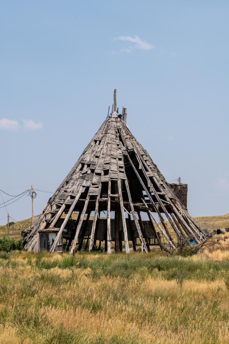 Une grande structure en bois patinée par les intempéries, en forme de tipi, se dresse dans un champ d’herbe sèche sous un ciel bleu limpide. L’ossature en bois de la structure est en grande partie apparente; de nombreux bardeaux manquant ou tombant, laissant apparaître le squelette nervuré en dessous. Visibilité masquée.