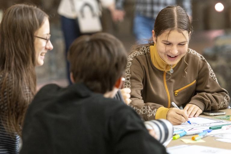 Deux adolescentes sont assises à une table. Elles rient et colorient un dessin avec des marqueurs, tandis qu’une autre personne est assise à proximité, dos à la caméra. Visibilité masquée.